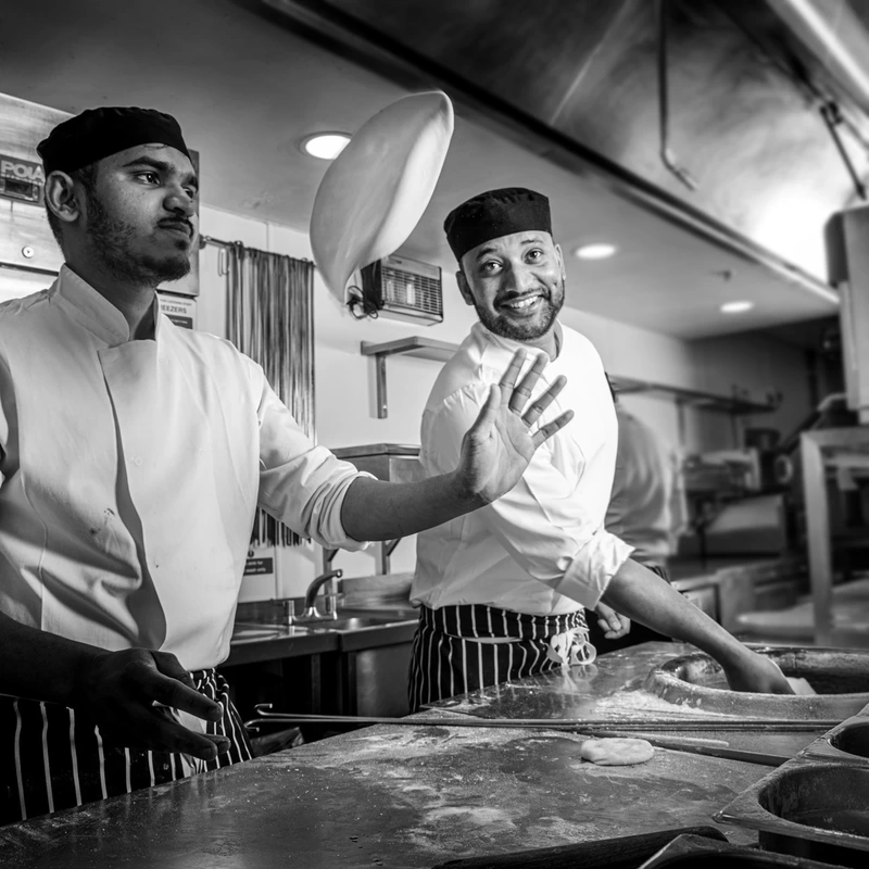 2 chefs making naans in the kitchen. One throwing a naan and one smiling