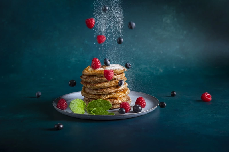 Raspberries and blueberries being dropped on a pile of pancakes