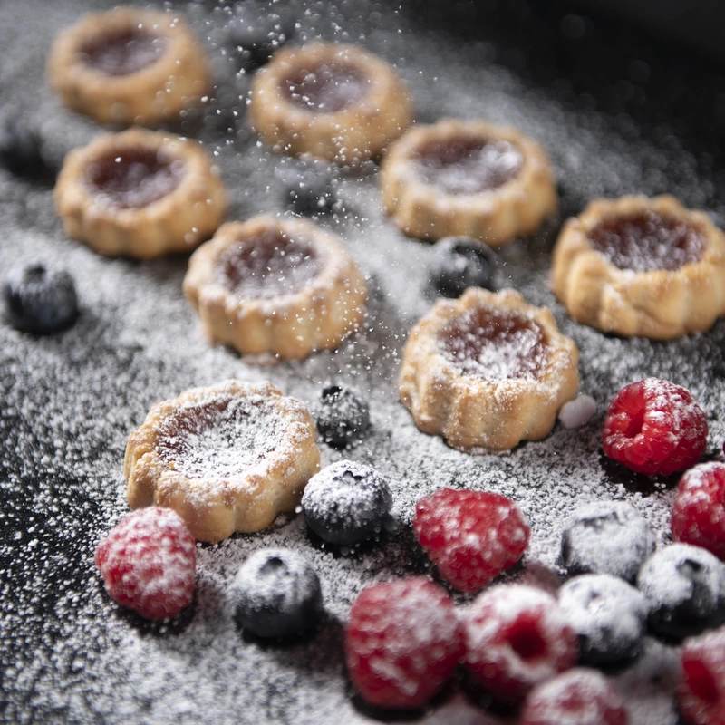Tarts and fresh berries on black tray dusted in icing sugar